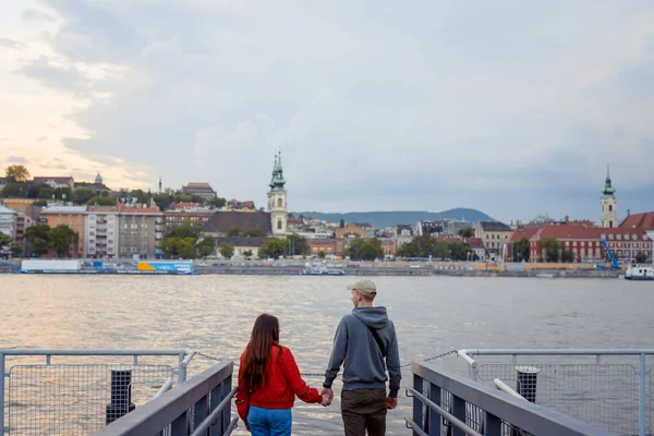Zadní pohled atraktivní pár v lásce se drží za ruce a užívat si krajinu řeky Dunaj v Budapešti, Maďarsko během západu slunce. — Stock fotografie