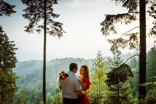 Счастливая влюбленная пара улыбается и нежно обнимается в лесу на фоне зеленых гор во время заката . — стоковое фото