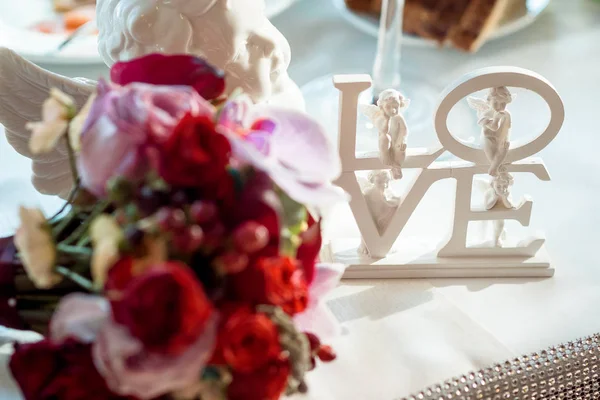 Close-up beeld van de witte letters liefde versierd met engelen geplaatst op de tafel in de buurt van wazig boeket kleurrijke rozen. — Stockfoto