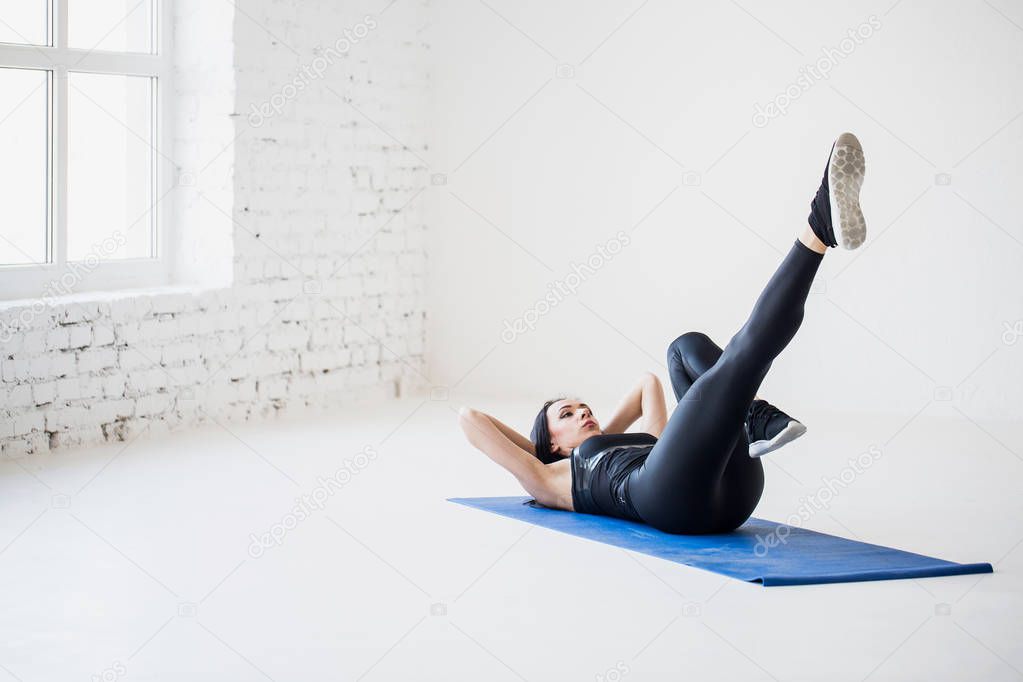 Back view of the sports lade warming up by lying on the exercise mat and doing bicycle crunches in the isolated white studio.
