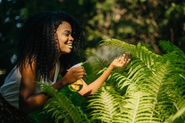 Güzel gülümseme, yeşil ruj ve göz gölgeler ferns için yeşil plastik tetikleyici sprey kullanırken dikkat çekici genç ve güzel kıvırcık Afrikalı kız portresi.