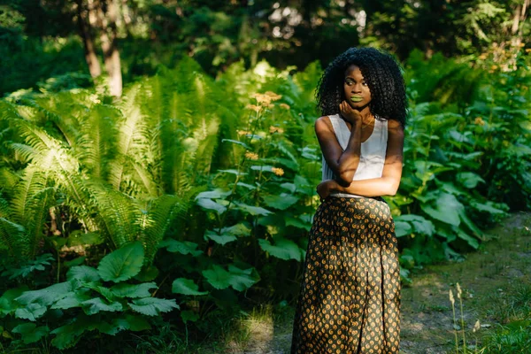 Retrato da jovem menina africana atraente com maquiagem verde em roupas elegantes olhando atenciosamente de lado enquanto posando no campo verde . — Fotografia de Stock