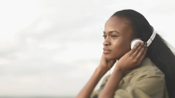 ナチュラルメイクで魅力的な若いアフリカの女の子の側の肖像画は屋外の白い背景でヘッドフォンで音楽を聴く. — ストック写真