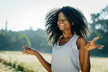Yeşil göz gölgeler ve kulaklık güneşli çayır üzerinde müzik keyfi güzel gülümseme ile büyüleyici genç Afrikalı kız portresi.