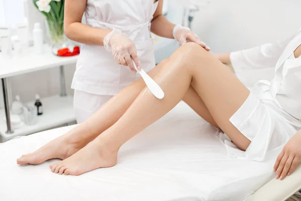 L'esthéticienne se prépare à l'épilation et applique la crème avec du bâton de cire sur les belles jambes féminines. Salon de beauté . — Photo