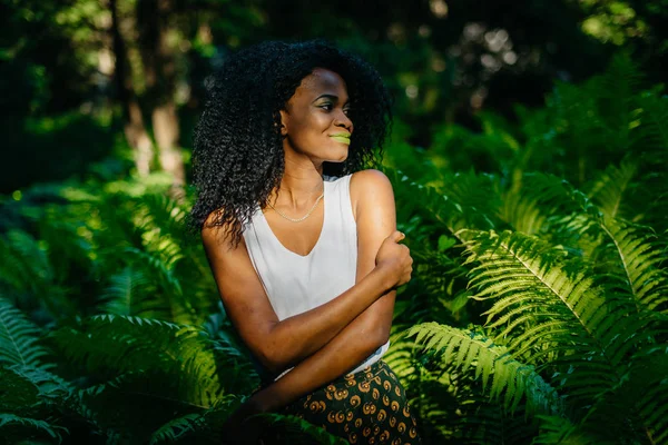 Retrato ao ar livre macio da linda menina africana com maquiagem verde pacificamente sorrindo e olhando para o lado enquanto estava entre os fetos . — Fotografia de Stock