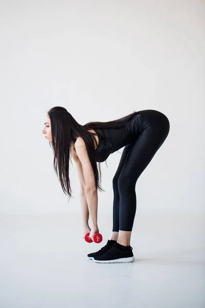 Die schlanke, muskulöse Brünette beugt sich mit den roten Hanteln im Studio. Seitenansicht. — Stockfoto