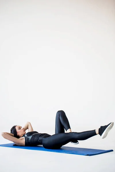 Спортивные занятия для начинающих. Молодая фитнес-девушка разогревается, делая велосипедные крэнчи на коврике для йоги в белой студии. . — стоковое фото