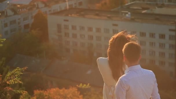 Vista posterior del hombre que viene a la joven morena y abrazándose mientras está de pie en el techo durante el atardecer. Vista panorámica . — Vídeo de stock