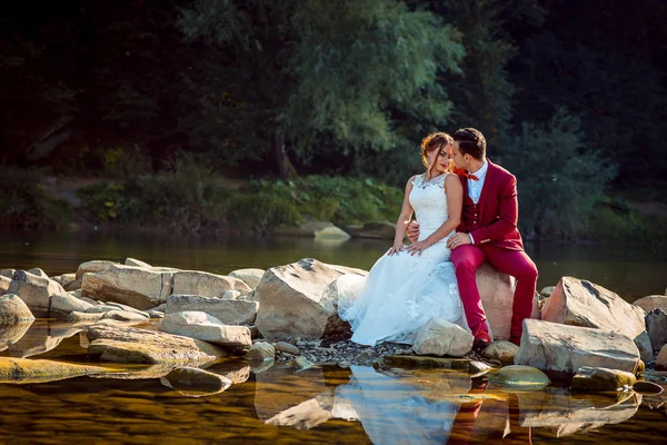 Romántico retrato de boda al aire libre de la hermosa pareja recién casada amorosa abrazándose mientras está sentado en la piedra cerca del río . — Foto de Stock