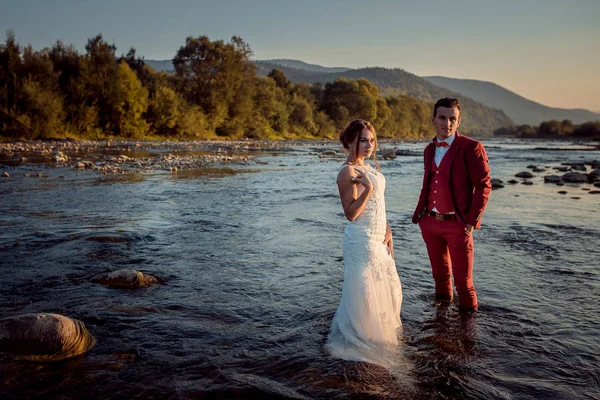 Retrato de la encantadora pareja de recién casados en el río al fondo del hermoso paisaje durante la puesta del sol . — Foto de Stock