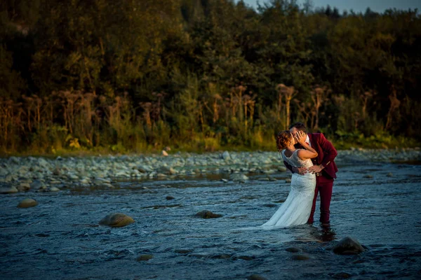 Romántico retrato al aire libre de la hermosa pareja alegre recién casada besándose en el medio del río durante el atardecer . — Foto de Stock