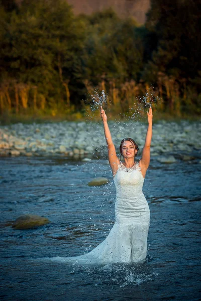 Encantadora noiva cabeça de gengibre no vestido de noiva está salpicando água enquanto está no meio do rio durante o pôr do sol . — Fotografia de Stock