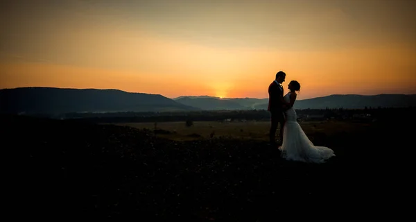 Tiro romântico dos belos recém-casados abraçando na borda das montanhas durante o pôr do sol . — Fotografia de Stock