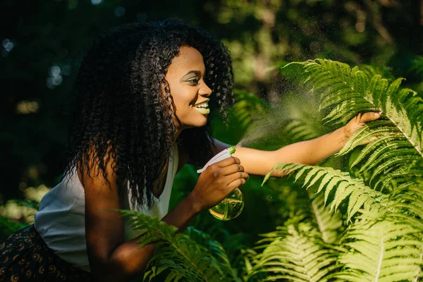 Atraktivní usměvavá Africká dívka s zelené oční stíny a rtěnku používá zelený plastový spouštěcí sprej pro kapradiny venkovní. — Stock fotografie