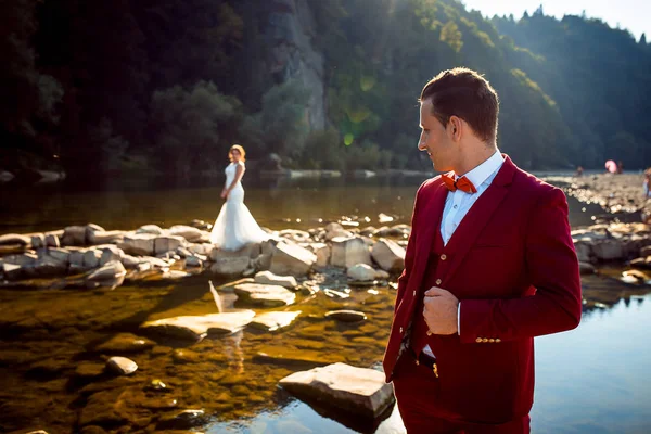 Крупним планом знімок красивого нареченого в червоному костюмі, який дивиться через плече на чарівну наречену, що стоїть далеко на каменях посеред річки. Красивий пейзаж . — стокове фото