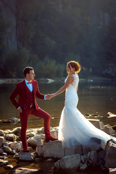 Повнометражний весільний портрет стильно одягненої молодої пари. Наречений тримає руки своєї чарівної імбирної нареченої, що стоїть на річкових каменях . — стокове фото