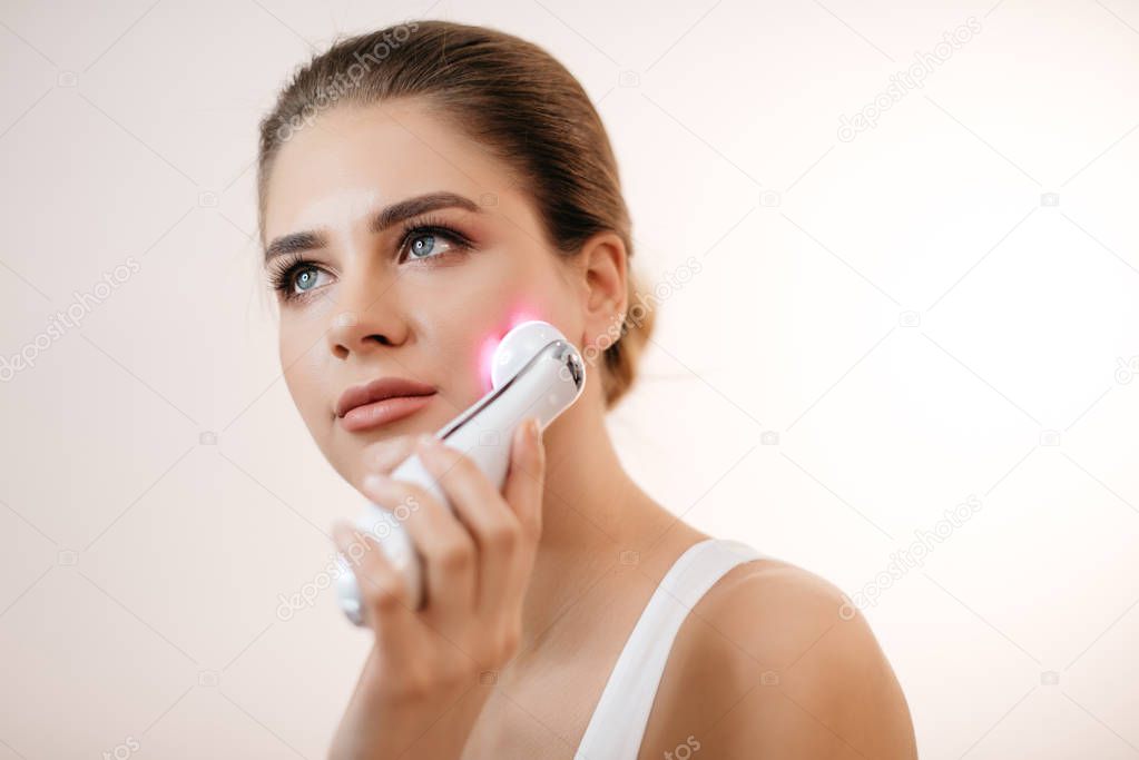 Young healthy fresh woman with natural make-up using the electric facial massager at isolated white background.