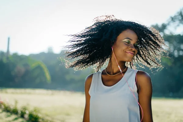 A jovem menina africana encantadora com sombras verdes e sorriso bonito está emocionalmente sacudindo seu cabelo encaracolado escuro enquanto ouve música em seus fones de ouvido no campo ensolarado . — Fotografia de Stock