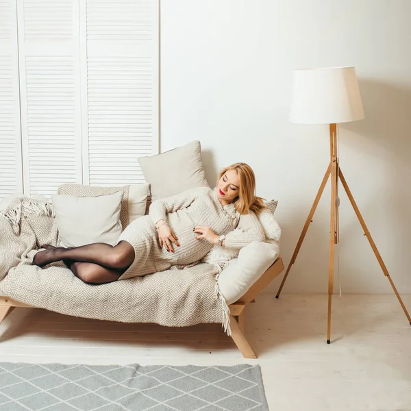 Beautiful blonde pregnant woman with red lipstick is touching her stomach and laying on the cozy sofa in the white room. — Stock Photo, Image