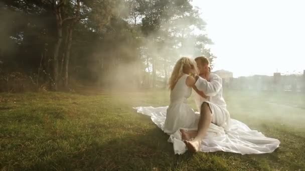 Fröhliches Liebespaar in Weiß küsst sich, während es im herbstlichen Park auf dem Rasen sitzt. — Stockvideo