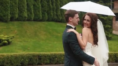 Genç çekici gülümseyen çift ayakta yağmur şemsiyesi altında komik portresi. Damat yavaşça onun sevgilisi ısırıyor.