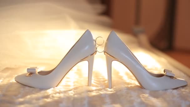 Datailní pohled zásnubní prsteny umístěna mezi bílá nevěsta highheels ležící na posteli. Pohyblivé sluneční světlo v pozadí. — Stock video