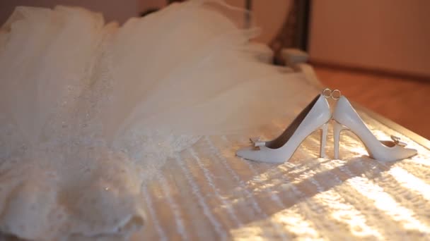 Närbild av vigselringar mellan bruden highheel liggande nära klänningen på sängen. Det rörliga solljuset. — Stockvideo