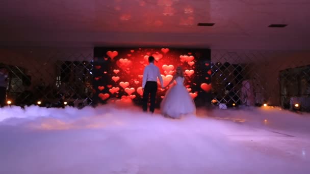 The beautiful happy couple of newlyweds are dancing their first dance at the background of the screen display with hearts and are shrouded by the white fume. Restaurant location. — Stock Video