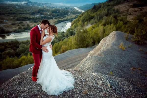 Feliz casal recém-casado é ternamente abraçando na borda das montanhas no fundo da magnífica paisagem do rio e florestas . — Fotografia de Stock