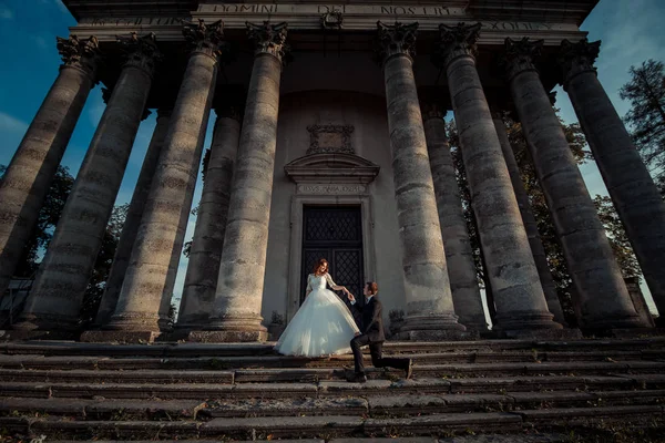 Ritratto di nozze all'aperto degli splendidi sposi. Lo sposo bello sta tenendo la mano della sposa affascinante mentre in piedi su un ginocchio sulle scale della vecchia chiesa barocca . — Foto Stock