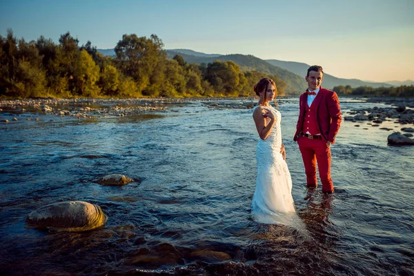 Atractiva pareja de recién casados en el río. El fondo de los bosques y montañas durante la puesta del sol . — Foto de Stock