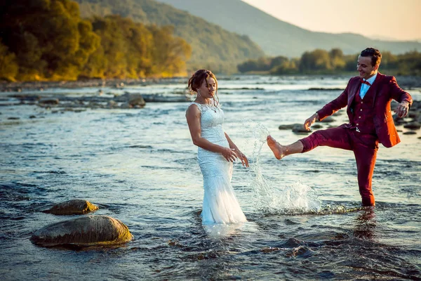 Feliz lindo casal recém-casado se divertindo na água, enquanto o noivo está salpicando água em sua linda noiva. Bela paisagem durante o pôr do sol . — Fotografia de Stock