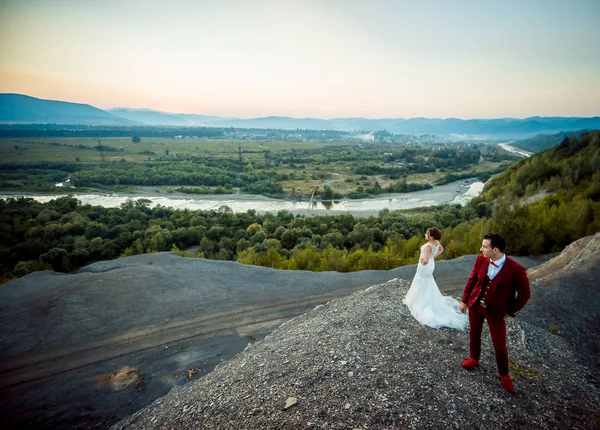 Encantador casal recém-casado na rocha está desfrutando a bela paisagem de rios, montanhas e florestas . — Fotografia de Stock