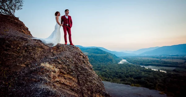 Modne newlywed para trzymając się za ręce na skraju skały. Piękny krajobraz, rzeki, lasy i góry podczas zachodu słońca. — Zdjęcie stockowe