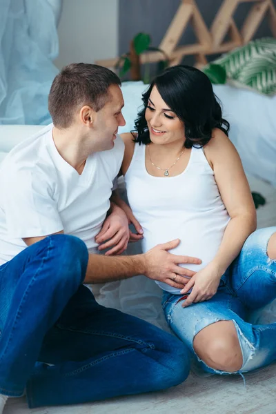 Портрет красивой молодой семьи, сидящей на полу. Красивый муж гладит живот своей прекрасной беременной жены . — стоковое фото