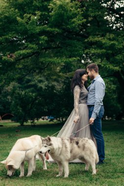 Sensitive portrait of the happy elegant charming couple rubbing noses during their walk with two huskies in the park. The attractive brunette in the long evening dress. clipart