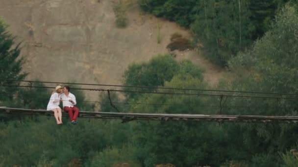 La feliz pareja de viajeros se besan mientras están sentados en el puente colgante de madera que cruza el río. Vista del dron . — Vídeo de stock