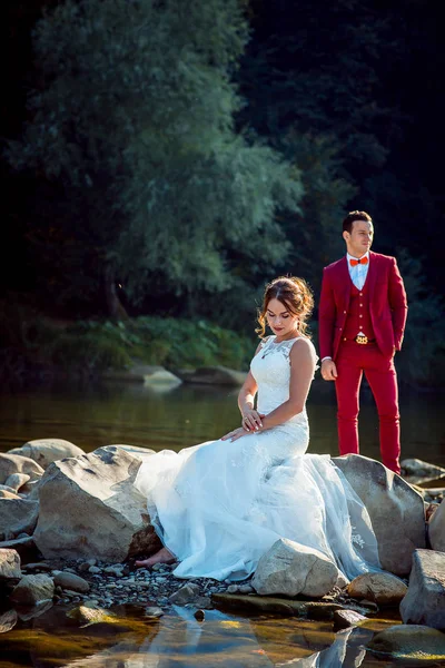 Свадебный наружный портрет. Привлекательная рыжая невеста сидит на камне у реки, а жених в красном стоит позади . — стоковое фото