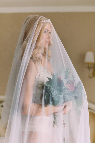Блондинка невеста с длинной вуали одета в сексуальное белое белье стоя на кровати и держа свадебный букет — стоковое фото
