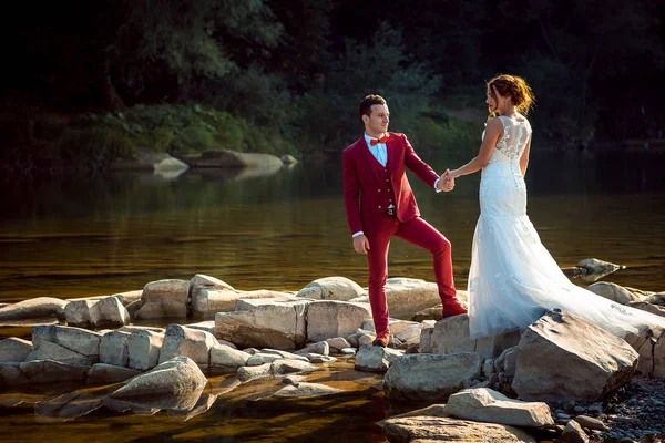 Горизонтальний знімок стильного нареченого в червоному костюмі, що тримає руки своєї нареченої на камені посеред річки . — стокове фото