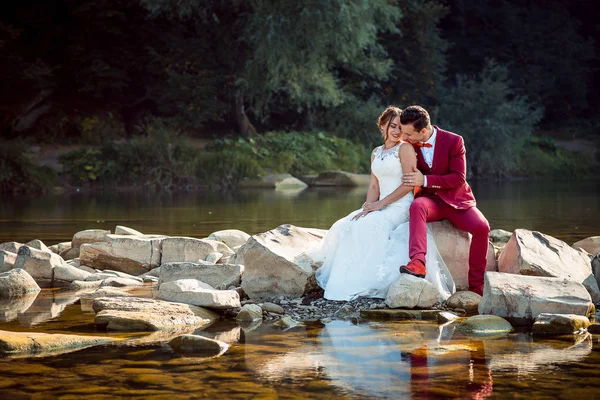 Горизонтальний знімок веселої чарівної новоспеченої пари, яка сміється і сидить на щілинах біля красивої річки . — стокове фото