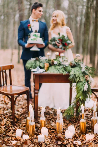 Hermosa pareja de boda posando con pastel en las manos cerca de la mesa vintage. Fondo del bosque otoñal — Foto de Stock