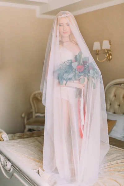 Blonde bride with long veil dressed in sexy white underwear standing on the bed and holding wedding bouquet