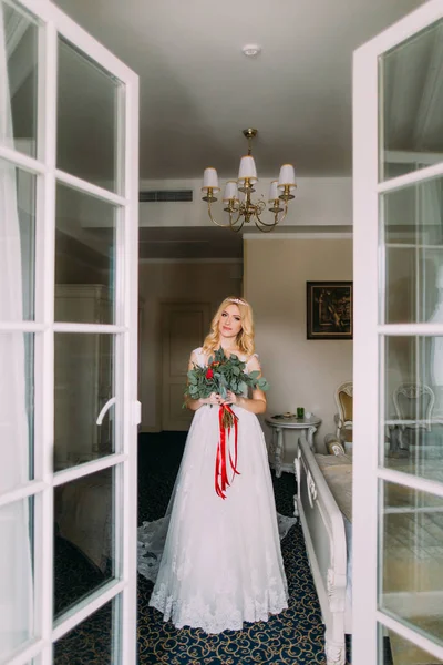 Belle mariée blonde avec bouquet de roses rouges debout près de la fenêtre — Photo