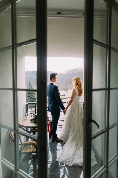 Casal de casamento encantador de pé e de mãos dadas na varanda. Visão traseira — Fotografia de Stock