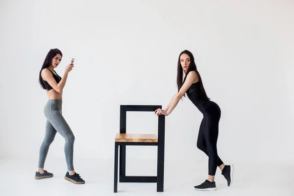 Full-length view of two charming athletic fitness women. One of them is taking photo of her friend leaning on the chair at the white background. — Stock Photo, Image