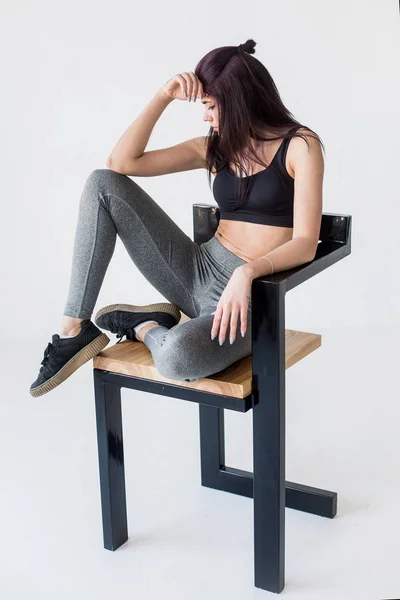 Die junge muskulöse Sportlerin posiert auf dem Stuhl und blickt zur Seite in das weiße Studio. Ansicht in voller Länge. — Stockfoto