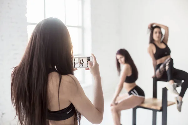 Rear view of the brunette taking photo of her two girl friends in short tops and shorts posing on the chair in white studio. Focus on the phone screen. — Stock Photo, Image