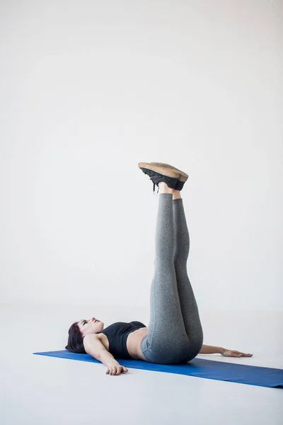 Pelatih wanita muda yang menarik adalah melakukan latihan kaki. Wanita atletik berbaring di tikar yoga dan mengangkat kakinya ke atas. Lokasi studio . — Stok Foto